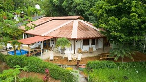 A house with a sloped roof, a swimming pool, and a wooden deck with outdoor furniture. - Paradiselodge - Poolvilla - next to Nauyaca (Platanillo de Baru)
