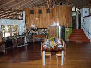 Una cocina amplia con paredes de madera, un comedor con una mesa larga dispuesta para una comida, y una escalera que conduce a un nivel superior.