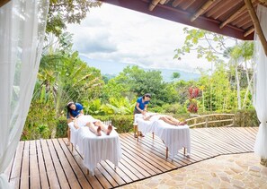 Terraza de madera con dos camillas de masaje, rodeada de vegetación exuberante y vistas a montañas distantes.