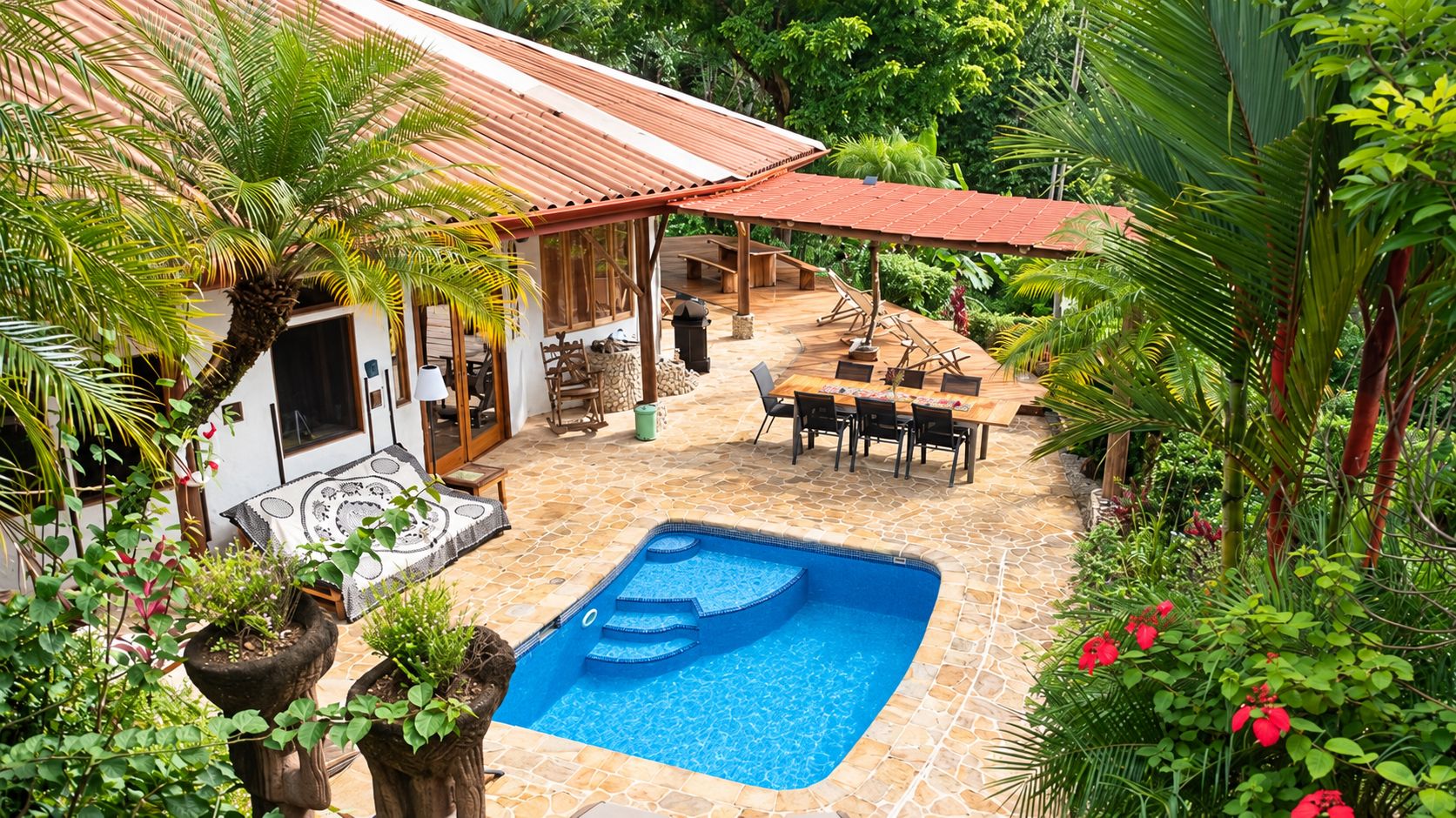 Un área de piscina con un deck de baldosas, una zona de estar cubierta y exuberante vegetación circundante.