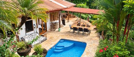 Un área de piscina con un deck de baldosas, una zona de estar cubierta y exuberante vegetación circundante.