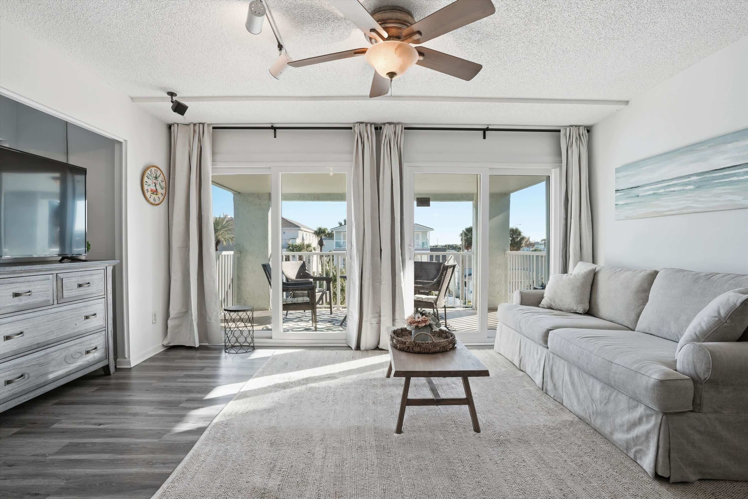 A spacious living room with a large sofa, a coffee table, and a television on a stand. There is a ceiling fan, a clock, and a view of the outside through large sliding doors.