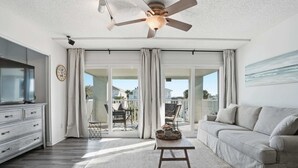 A spacious living room with a large sofa, a coffee table, and a television on a stand. There is a ceiling fan, a clock, and a view of the outside through large sliding doors.