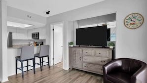 A modern living room with a flat-screen TV, a dark leather sofa, and a dining area with two bar stools.