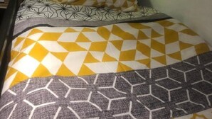 Un lit simple avec une couvre-lit à motifs jaune et blanc, un plaid géométrique gris et blanc, et un oreiller de couleur claire orné d’un motif géométrique.
