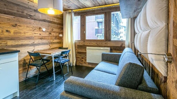 Une pièce chaleureuse avec un plafond en bois, un canapé, une table à manger avec des chaises et une fenêtre donnant sur des immeubles.
