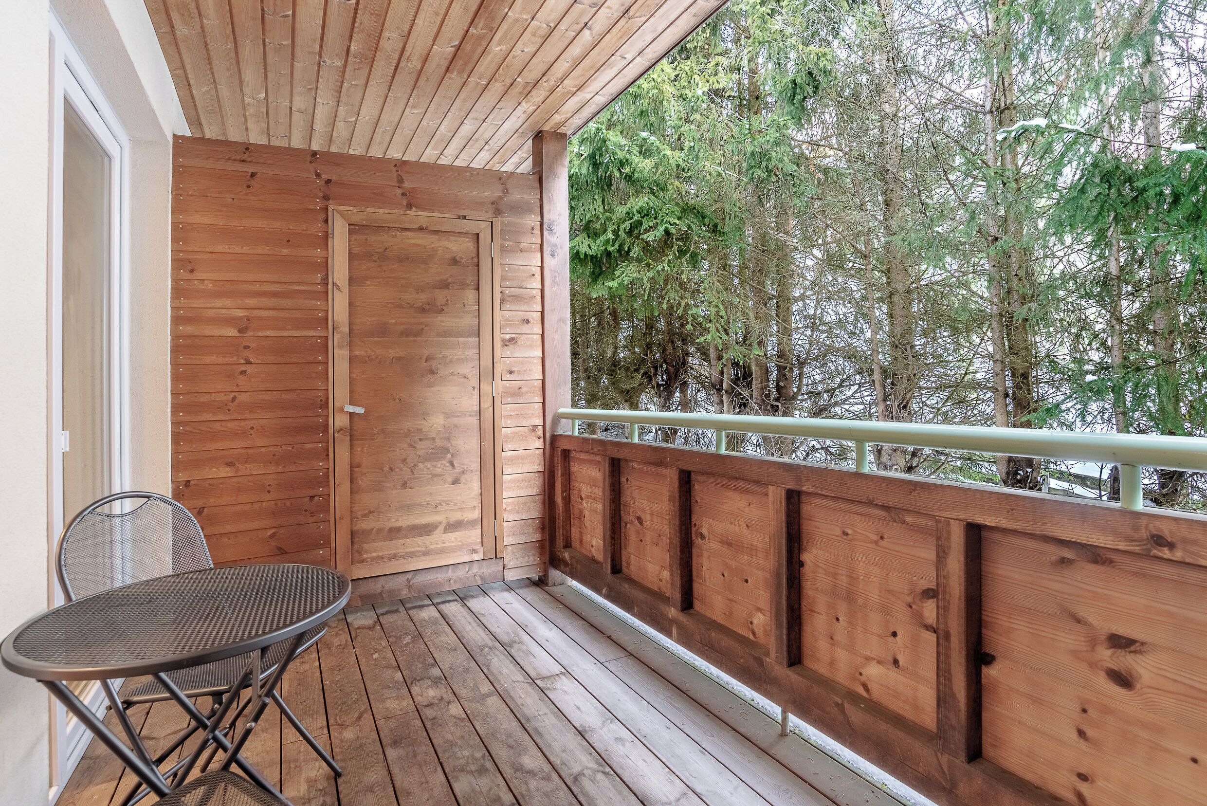 A wooden deck with a metal table and chair, a wooden door, and a view of trees.