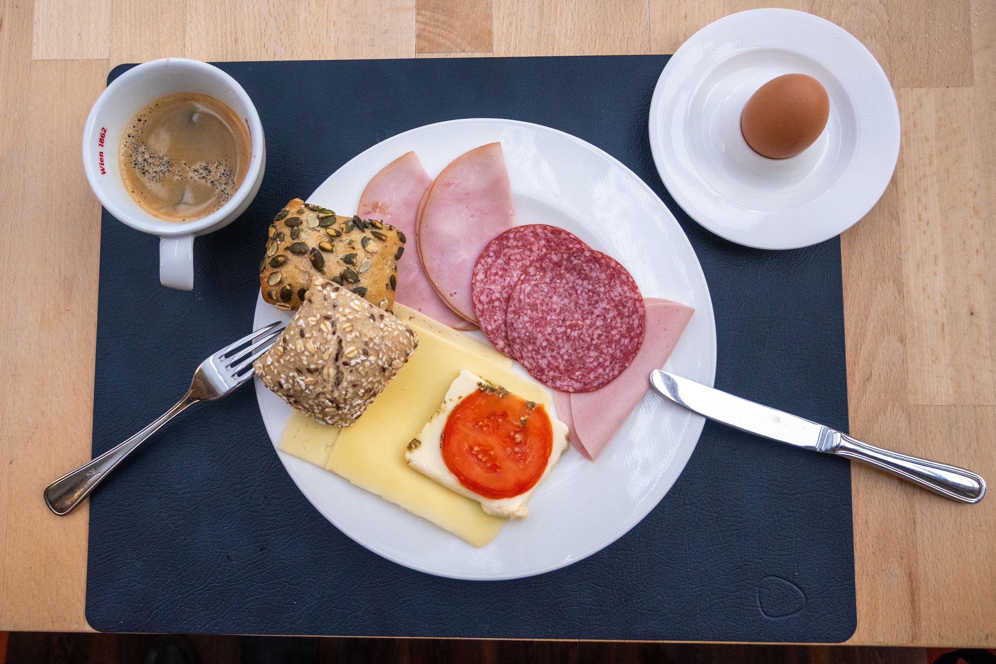 Een bord met ham, salami, kaas, tomaat en brood, een kop koffie en een mes op een tafel.