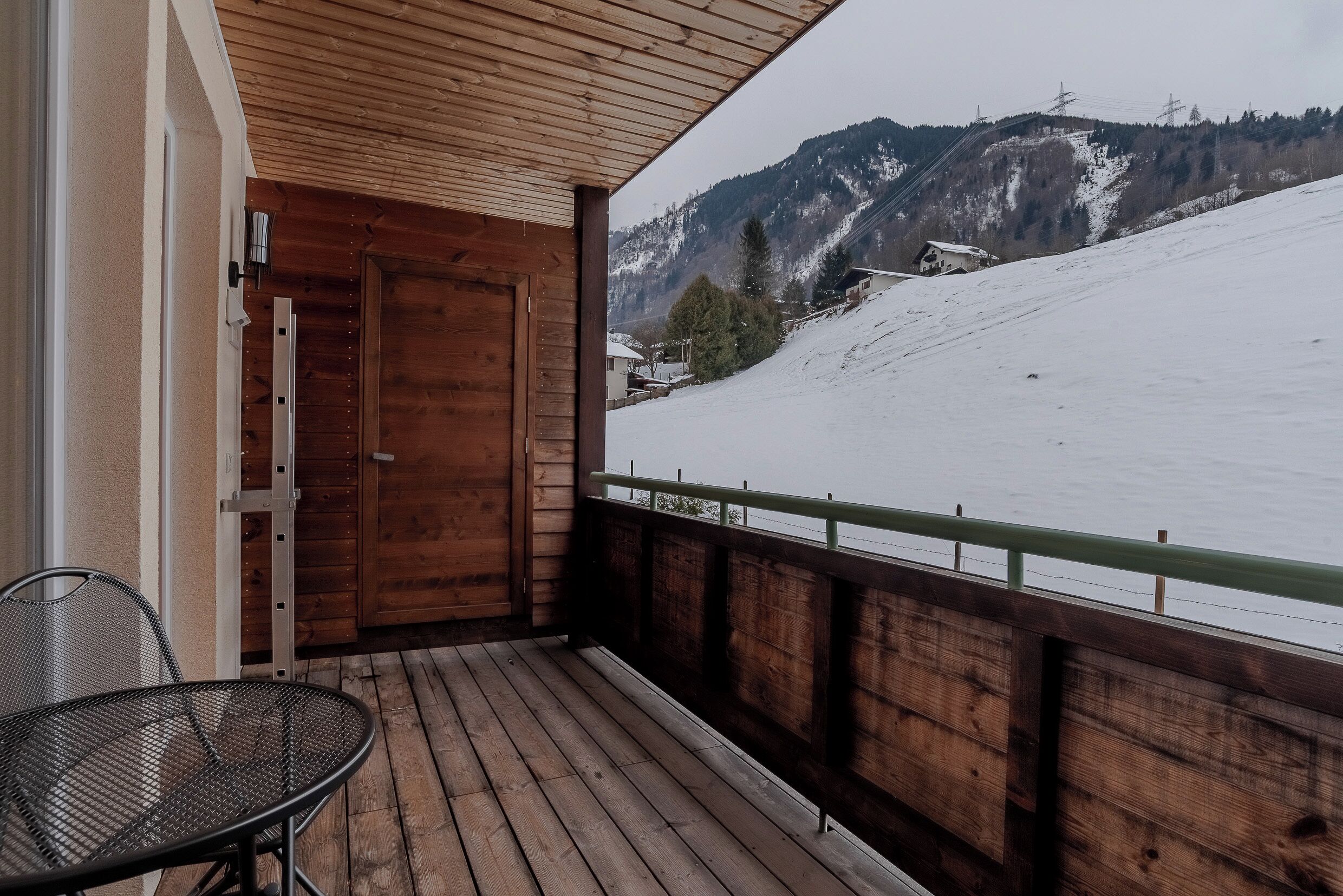 Una terraza de madera con una silla metálica, una puerta de madera y vistas a una montaña nevada.