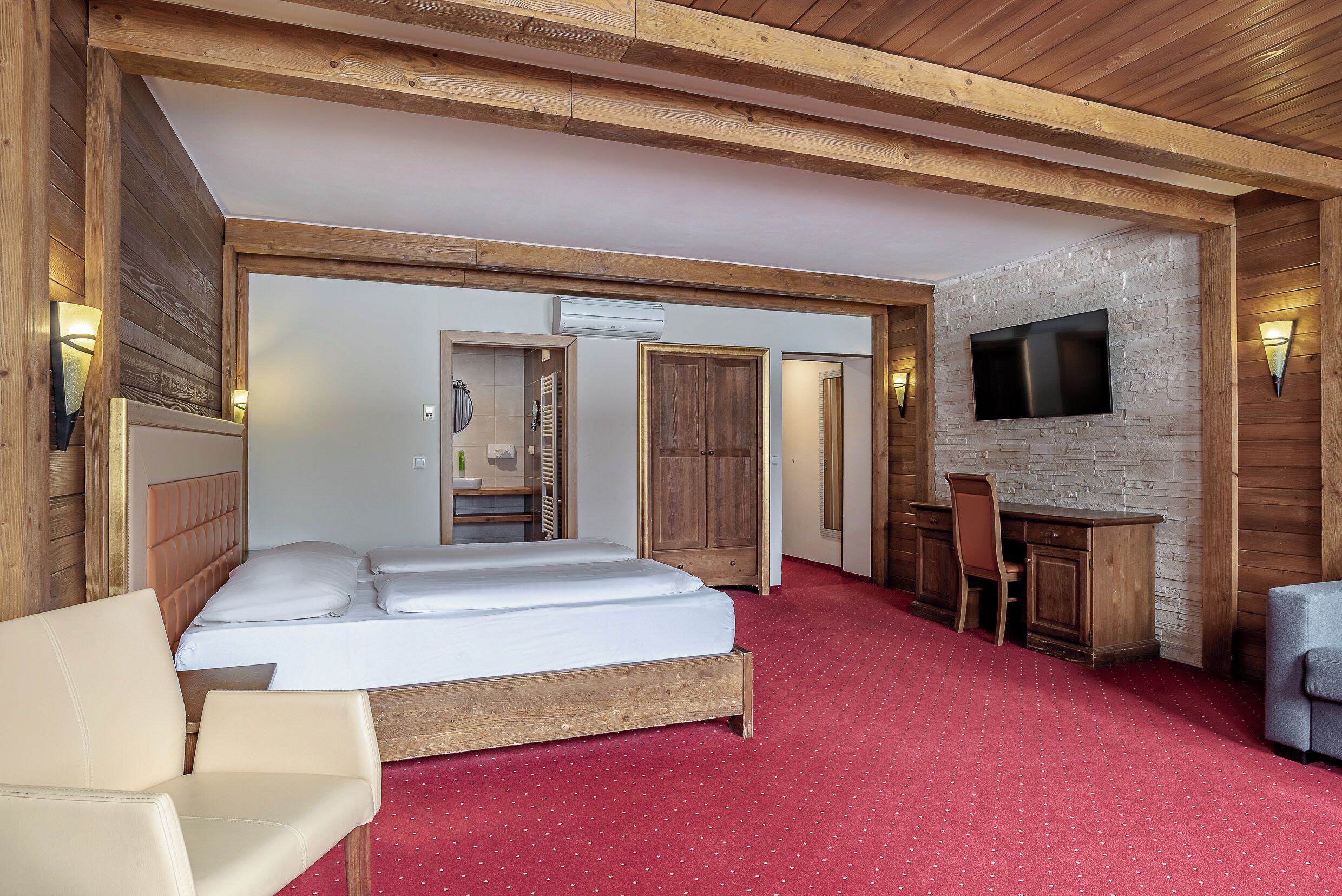A bedroom with a bed, a desk, a chair, a TV mounted on a wall, and a red carpet.