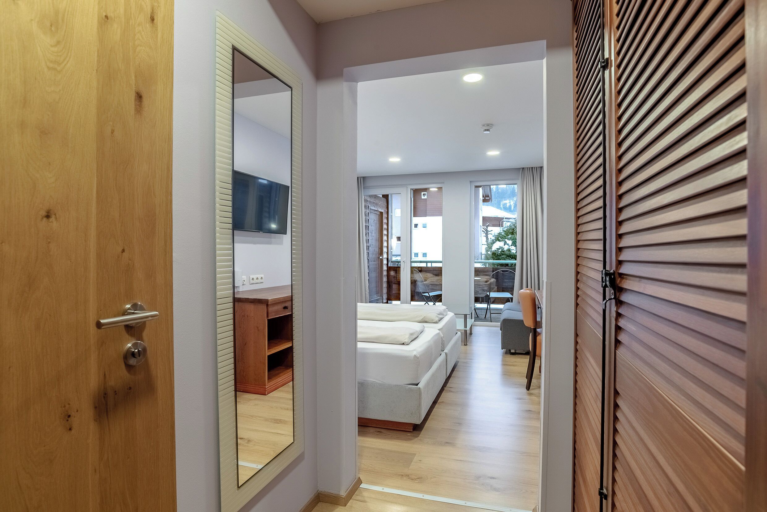 A hallway leading to a bedroom with a bed, a mirror, and a wooden door.
