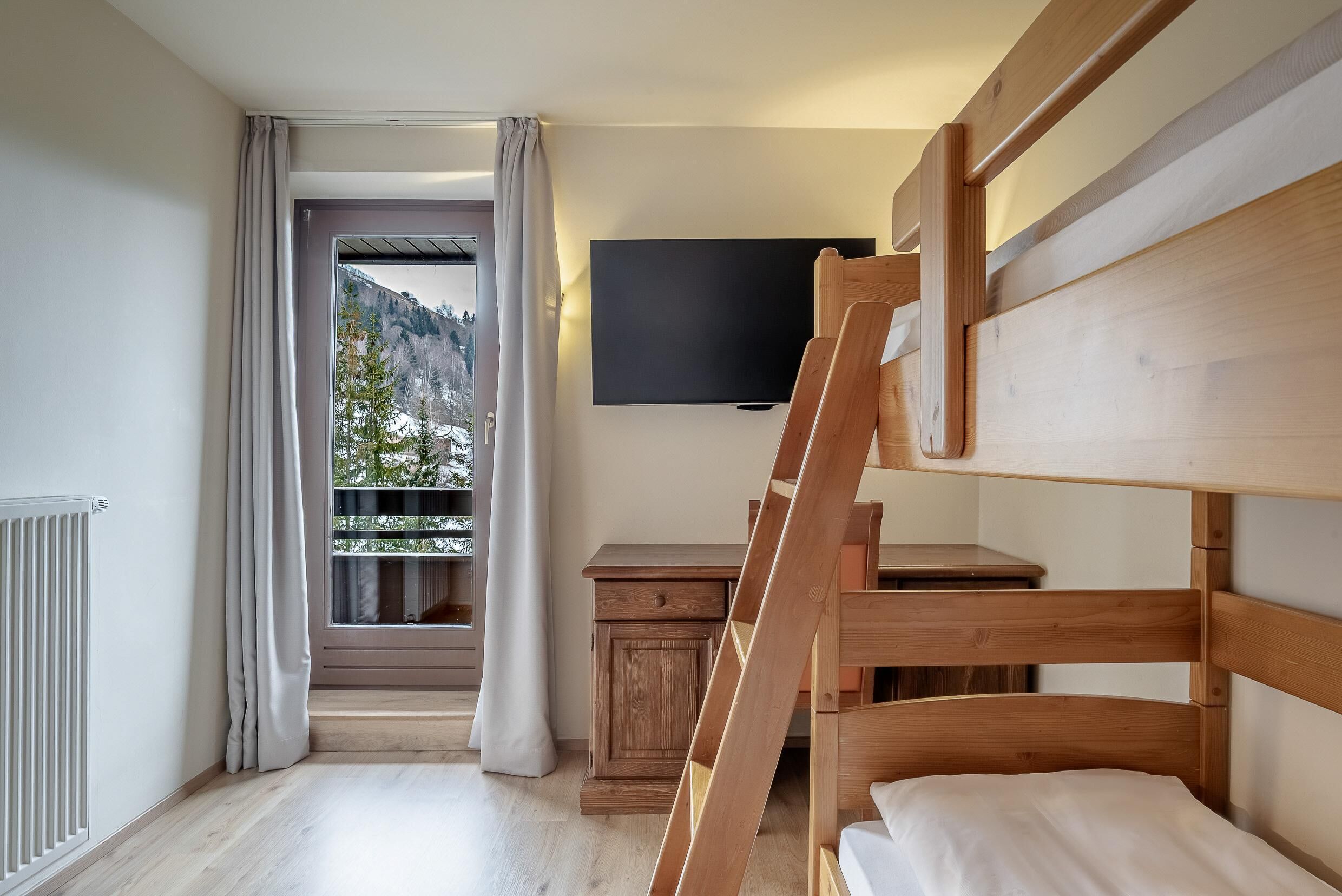 A bunk bed room with a TV, a small wooden desk, and a window with curtains.
