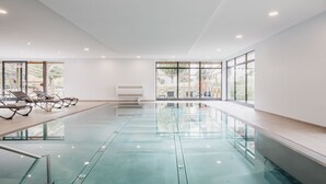 An indoor swimming pool with clear glass walls, a ceiling with recessed lighting, and a row of sun loungers along the left side.