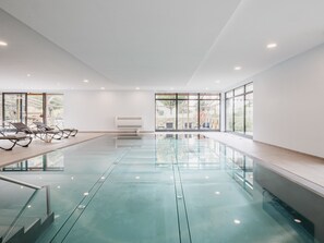An indoor swimming pool with clear glass walls, a ceiling with recessed lighting, and a row of sun loungers along the left side. - Hotel Victoriya (Kaprun)