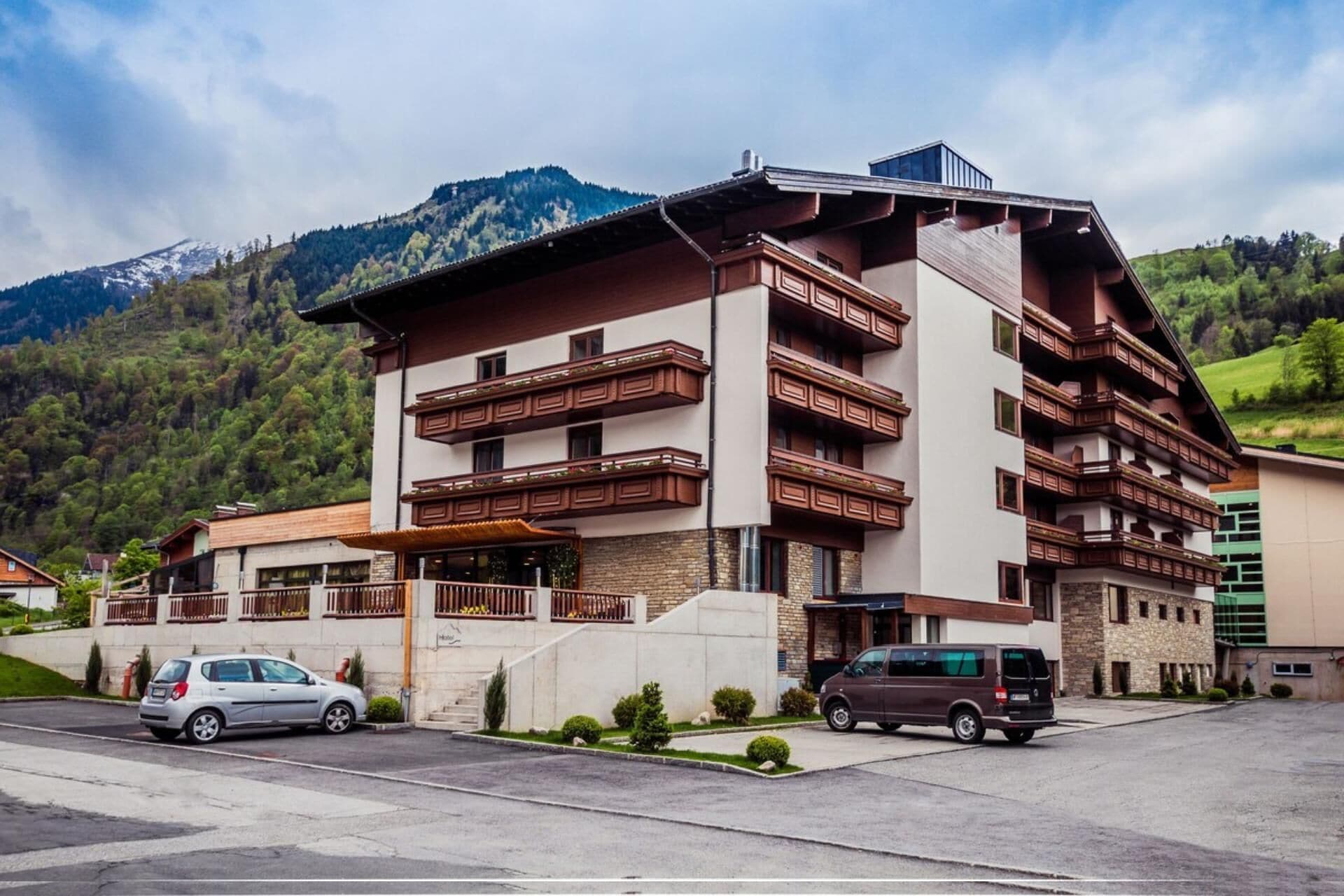 Hotel de diversos pisos amb balcons, zona d'aparcament i un paisatge de muntanyes al fons.