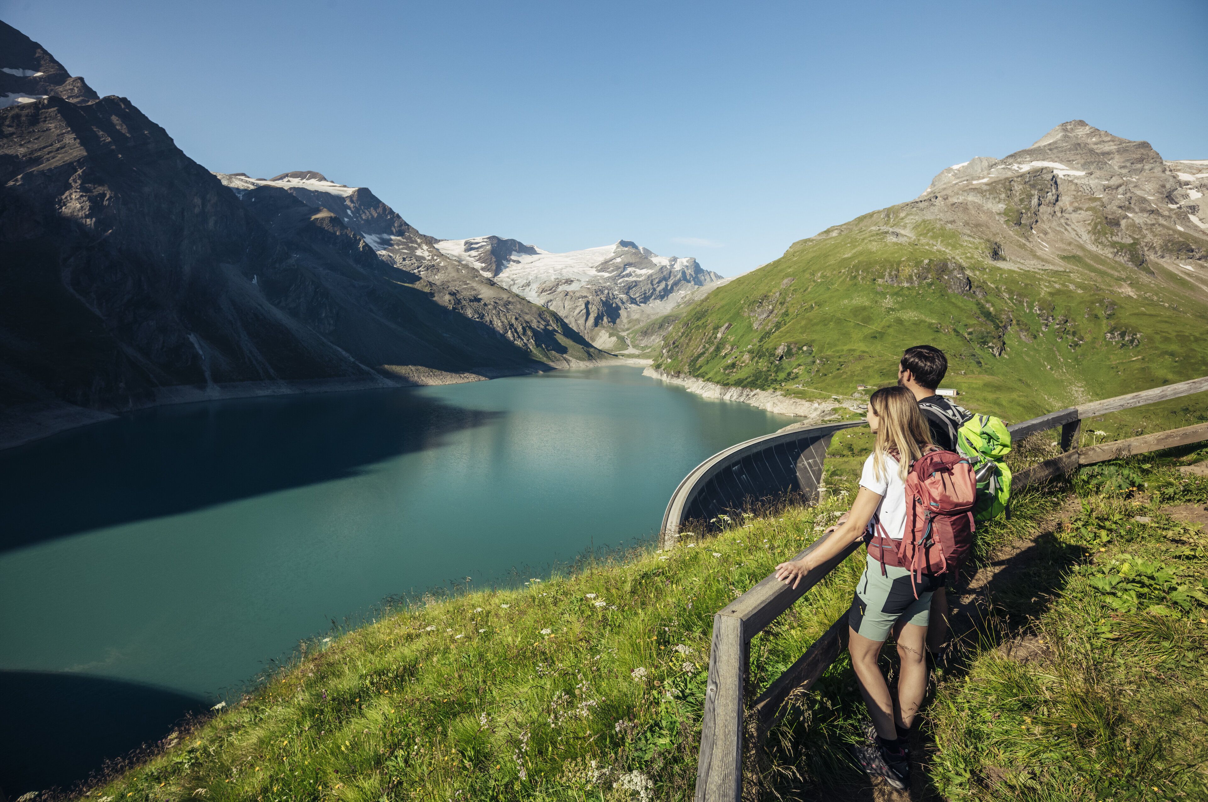 Eine Berglandschaft mit einem See, einer Holzbrücke und Wanderern mit Rucksäcken.