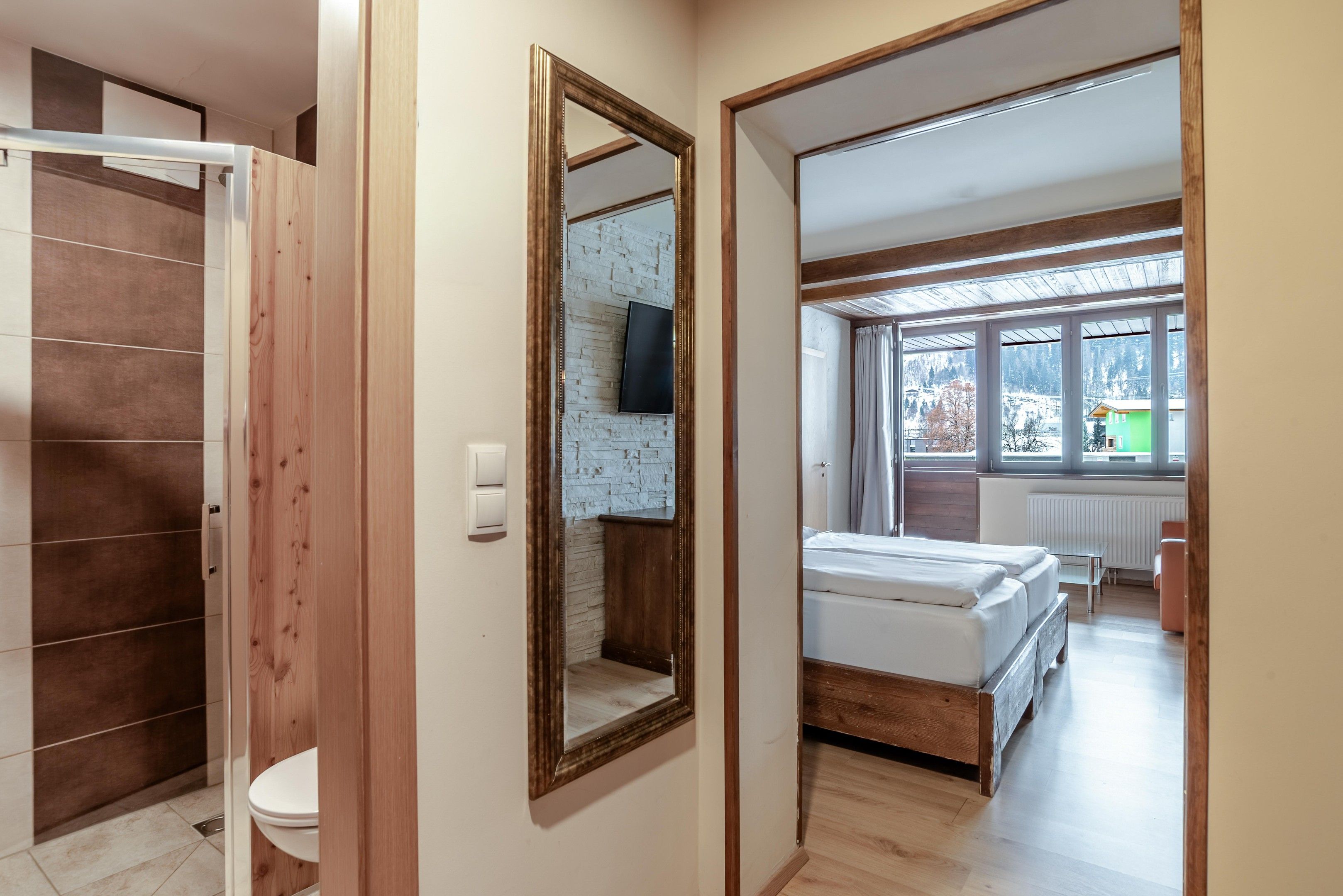 A hotel room with a wooden wardrobe, a mirror, a bed, a window with a view of a snowy landscape, and a mounted television.