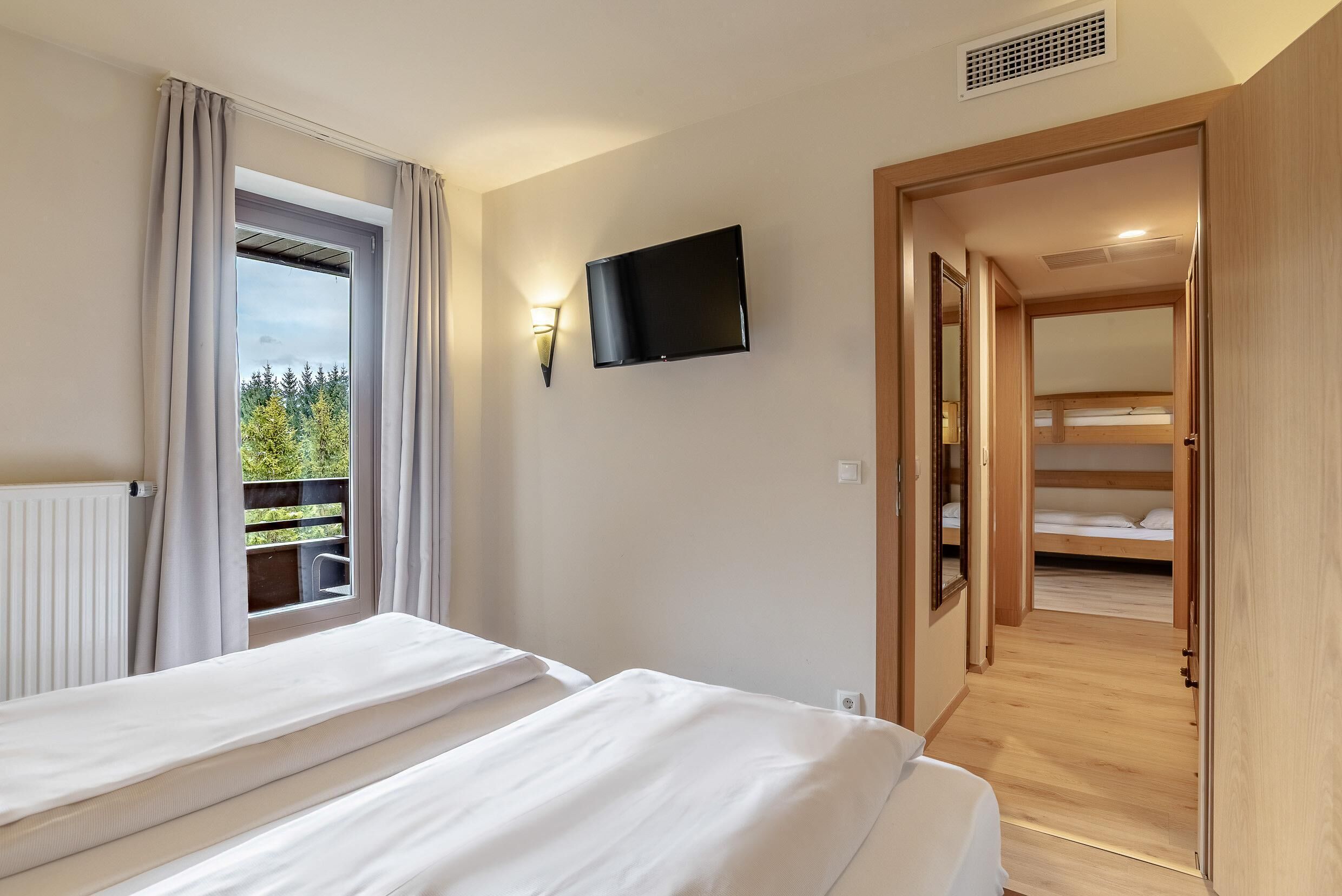 A room with two beds, a TV, a balcony with curtains, and a wooden door leading to another room.