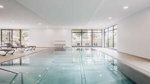 A modern indoor swimming pool with clear glass walls, sun loungers, and a view of the outdoors. - Hotel Victoriya (Kaprun)