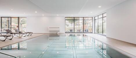 A modern indoor swimming pool with clear glass walls, sun loungers, and a view of the outdoors.