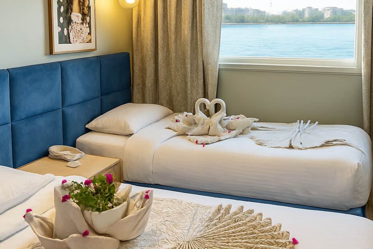 A hotel room with two beds, a blue headboard, a large window with a view of a body of water, and a framed picture on the wall.