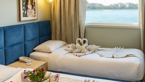 Une chambre d’hôtel avec deux lits, une tête de lit bleue, une grande fenêtre donnant sur un plan d’eau et un tableau encadré au mur.