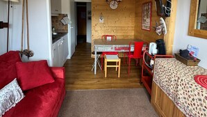Et kompakt værelse med tekøkken, spiseplads med bord og stole, seng med et mønstret dynebetræk og en rød sofa.