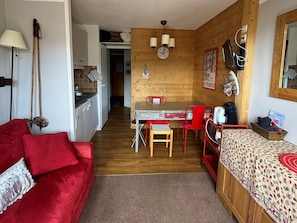 Ein kompakter Raum mit Küchenzeile, einer Essecke mit Tisch und Stühlen, einem Bett mit gemustertem Überwurf und einem roten Sofa.