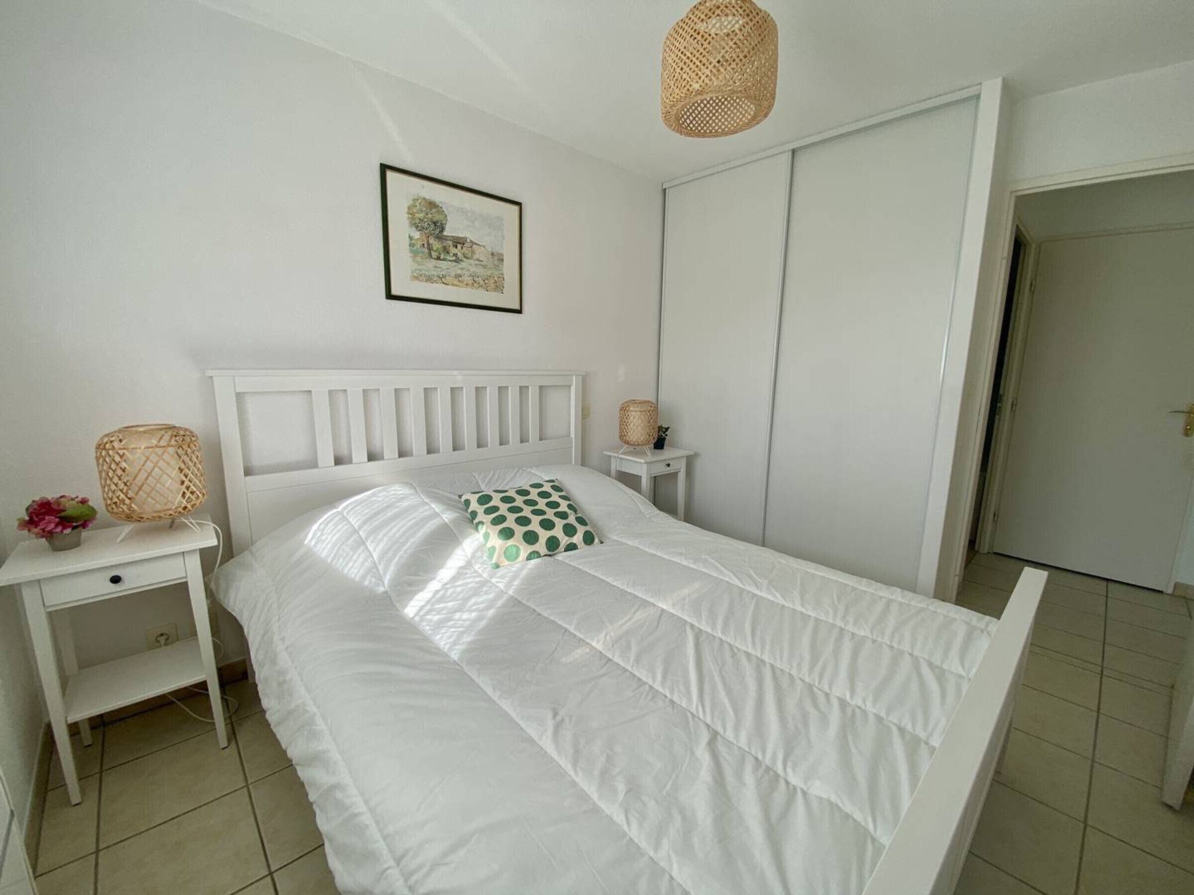 Une chambre à coucher bien rangée, avec un lit blanc, une table de chevet sur laquelle se trouve une lampe, et un tableau encadré accroché au mur.