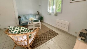 A living room with a sofa, a wicker chair, a small table with a vase, a window with curtains, and a radiator.