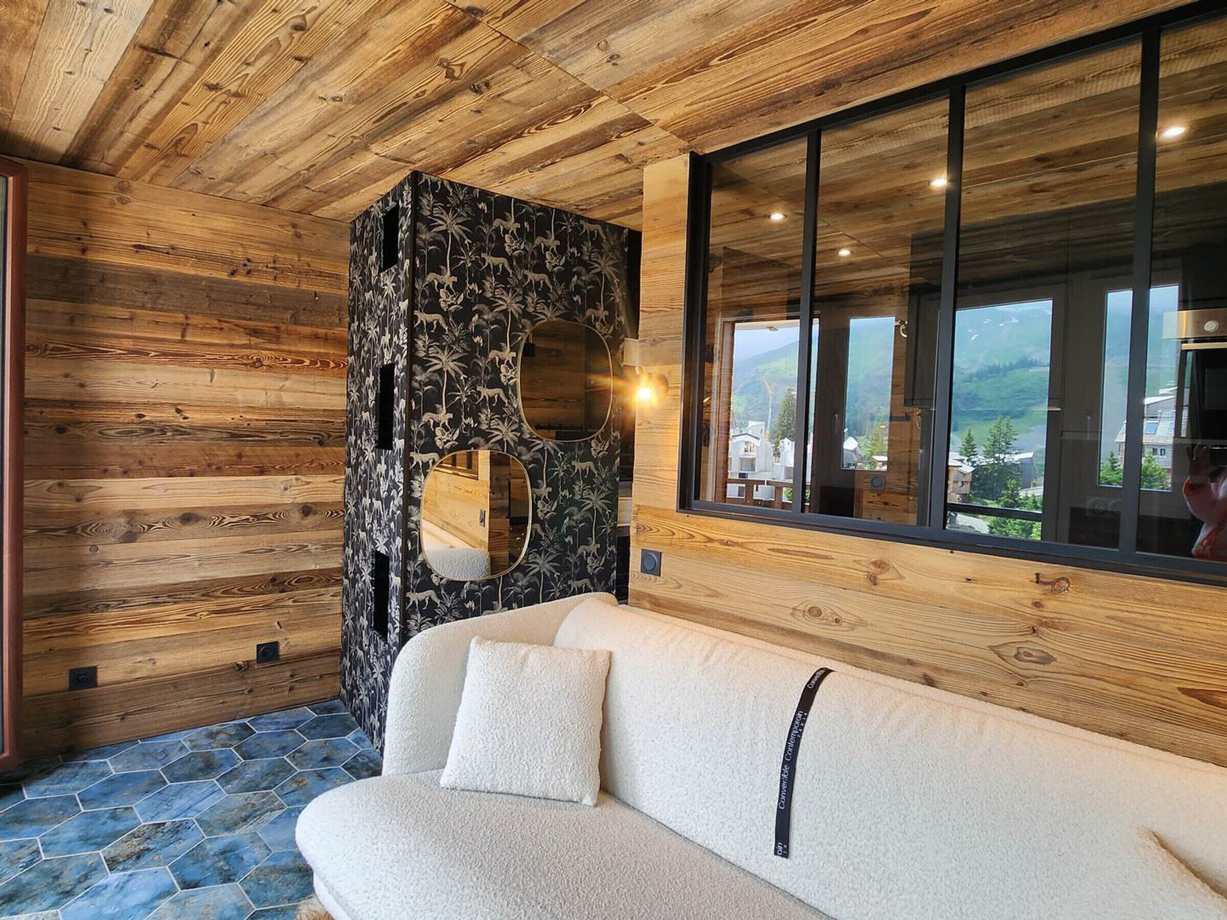 Een houten interieur met een bank, een decoratieve wand met een ronde spiegel, en een groot raam met uitzicht op de omgeving.