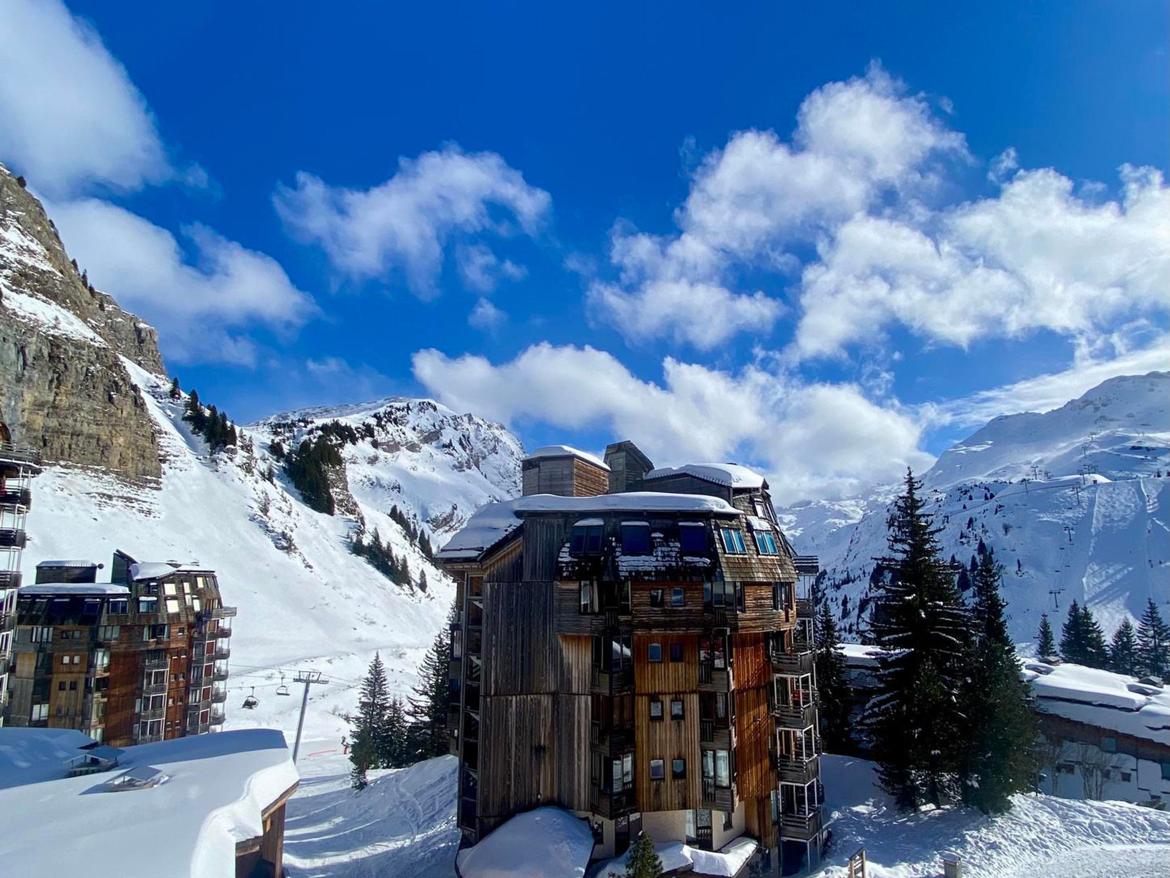 Eine Holzhütte in den Bergen mit mehreren Balkonen, umgeben von schneebedeckten Gipfeln und blauem Himmel.