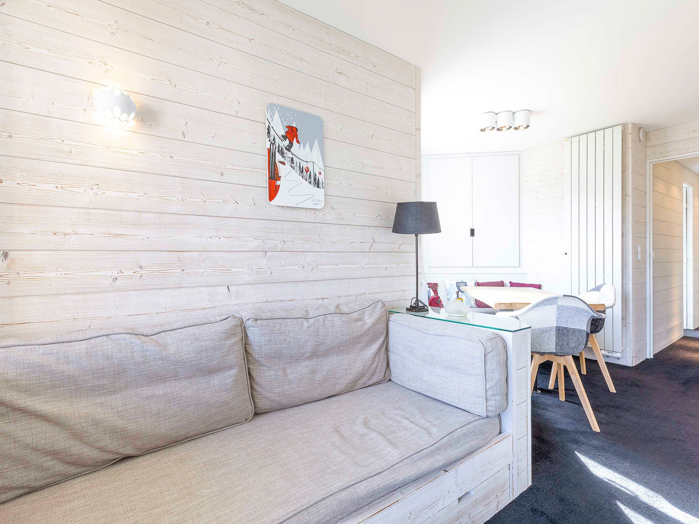 Ett rum med en ljus träpanel på väggen, en vit soffa, ett glasbord, en svart lampa och en matplats med ett vitt bord och stolar.