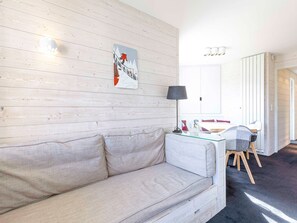 A room with a light-colored wooden wall, a white sofa, a glass coffee table, a black lamp, and a dining area with a white table and chairs.