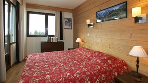 A bedroom with a wooden wall, a large bed with a red floral bedspread, two bedside lamps, a dresser, and a window with curtains. - 3-room apartment, sleeps 6 in Avoriaz (Avoriaz)