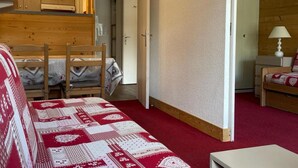 Una habitación pequeña con una cama nido de madera, una mesita con una silla, y una cama individual con colcha estampada de rojo y blanco.