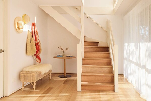 Una escalera que conduce a un piso superior, un banco de mimbre, una pequeña mesa redonda con libros y un florero, y sombreros en la pared.