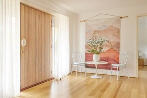 Una habitación con puerta de madera, cortinas blancas, una mesa redonda con dos sillas blancas y un cuadro abstracto grande en la pared.