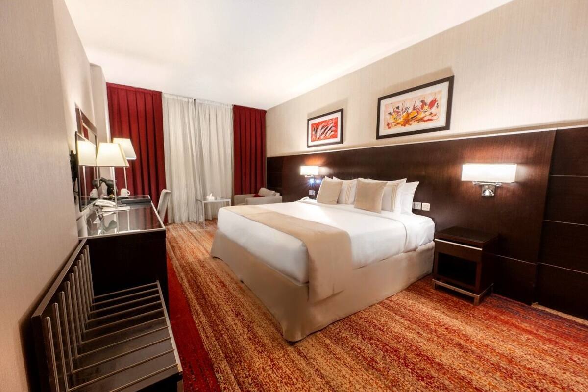 A hotel room with a large bed, a desk with a lamp, a chair, and a small table.