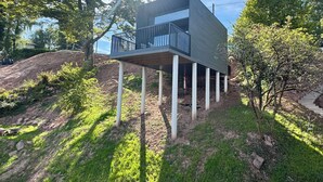 Ein modernes, zweistöckiges Haus mit Balkon, umgeben von Bäumen und einer grünen Böschung.