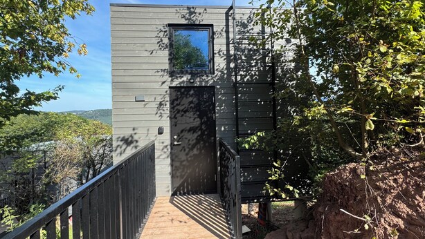 Ein modernes Gebäude mit Holzfassade, einem kleinen Fenster und einer Holztreppe, die zu einer Tür führt.