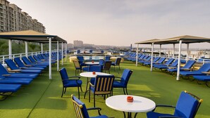 Eine Dachterrasse mit blauen Liegestühlen und Tischen, einem Boden aus grünem Kunstrasen und Blick auf Gebäude im Hintergrund.