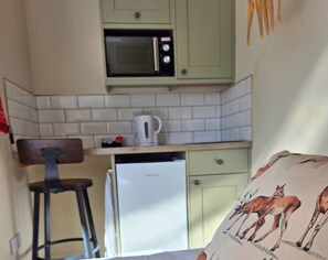A small kitchenette with a microwave, a coffee maker, and a refrigerator. There is a chair and a bed with a pillow featuring deer illustrations.