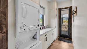 Een wasruimte met een voorlader wasmachine en droger, houten kasten en een deur naar een balkon met uitzicht op groen.