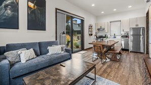 Een moderne woonkamer met een grijze bank, een houten salontafel en een eethoek met een houten tafel en stoelen. In de achtergrond is een keuken met roestvrijstalen apparatuur te zien.