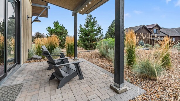 Een terras buiten met moderne lounge fauteuils, een houten pergola en een aangelegde tuin met diverse planten en bomen.
