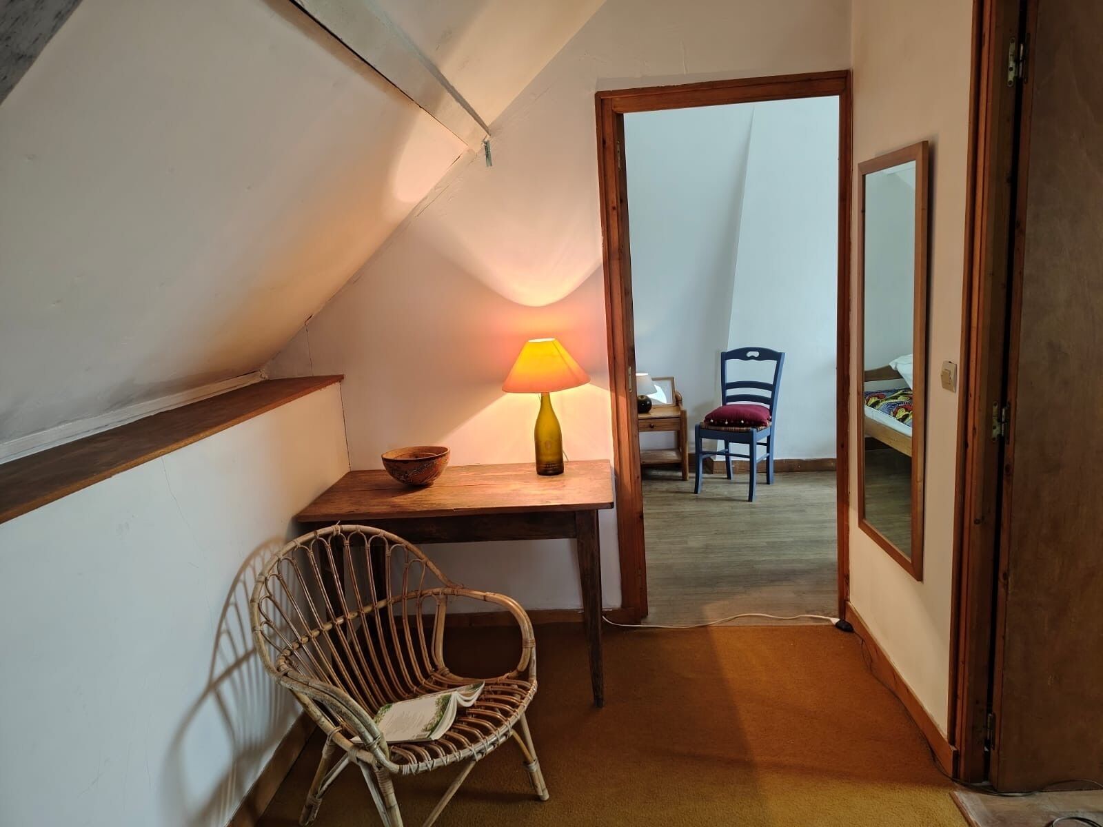 Habitació acollidora amb un sostre inclinat, una taula de fusta amb una làmpada, una cadira de vímet i un llit amb un llençol de colors.