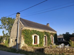 Una casa de piedra con muros cubiertos de hiedra, dos ventanas con contraventanas y un muro de piedra al frente.