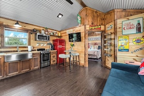 A rustic cabin-style kitchen with wooden walls, a red refrigerator, a stove, and a microwave. There is a bar area with stools and a television. - Hill Country Cabin - Cozy Rustic Getaway (Fredericksburg)
