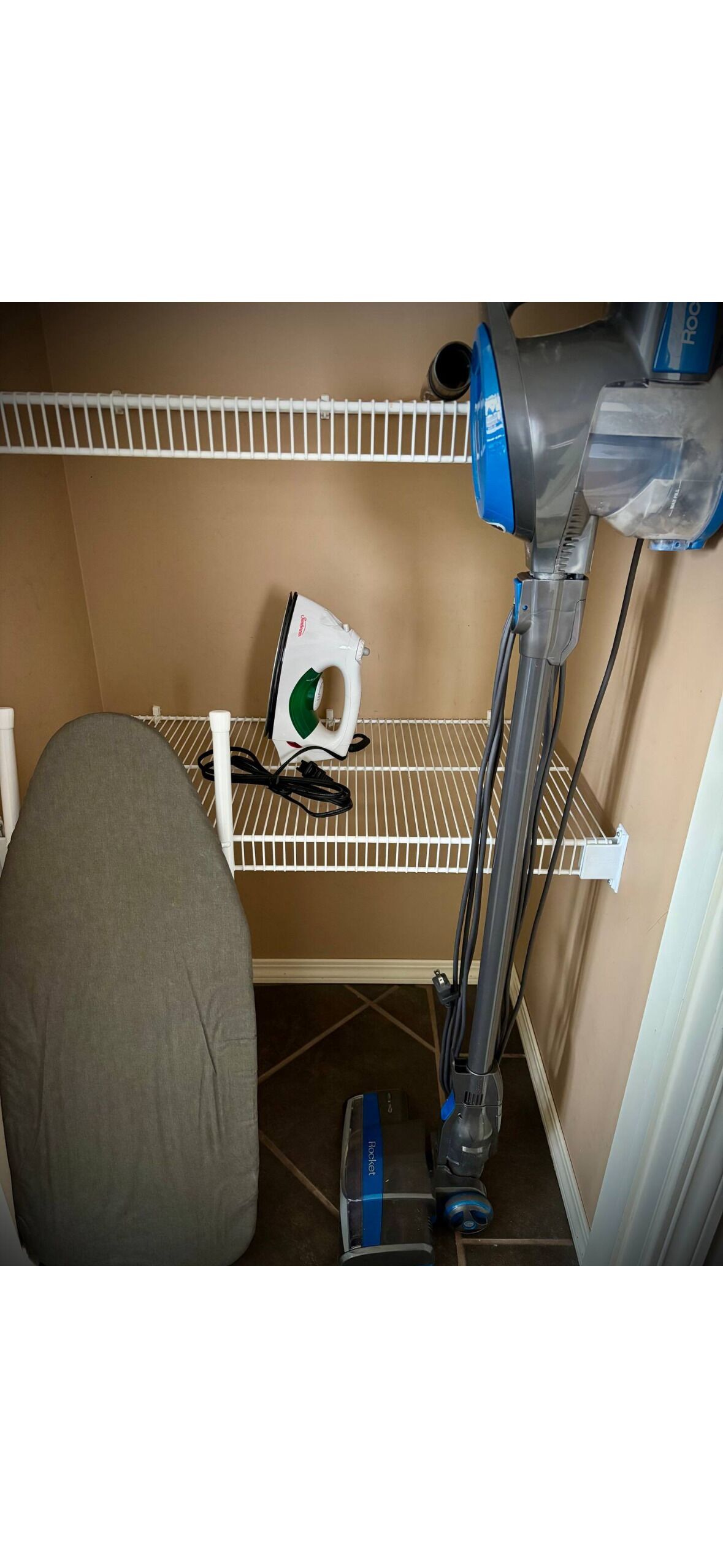 A compact storage area with a vacuum cleaner, an ironing board, and a shelf with a green iron.